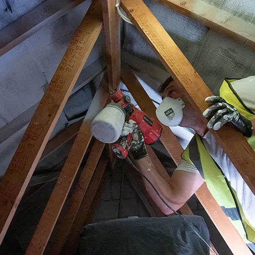 A construction worker wearing a high-visibility yellow vest and protective gloves uses a red electric nail gun with a white cup carrier to apply adhesive fasteners to wooden roof rafters.